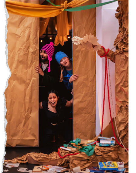 Three performers are peering through a gap in a wall looking inquisitive. There is a cardboard tree next to them