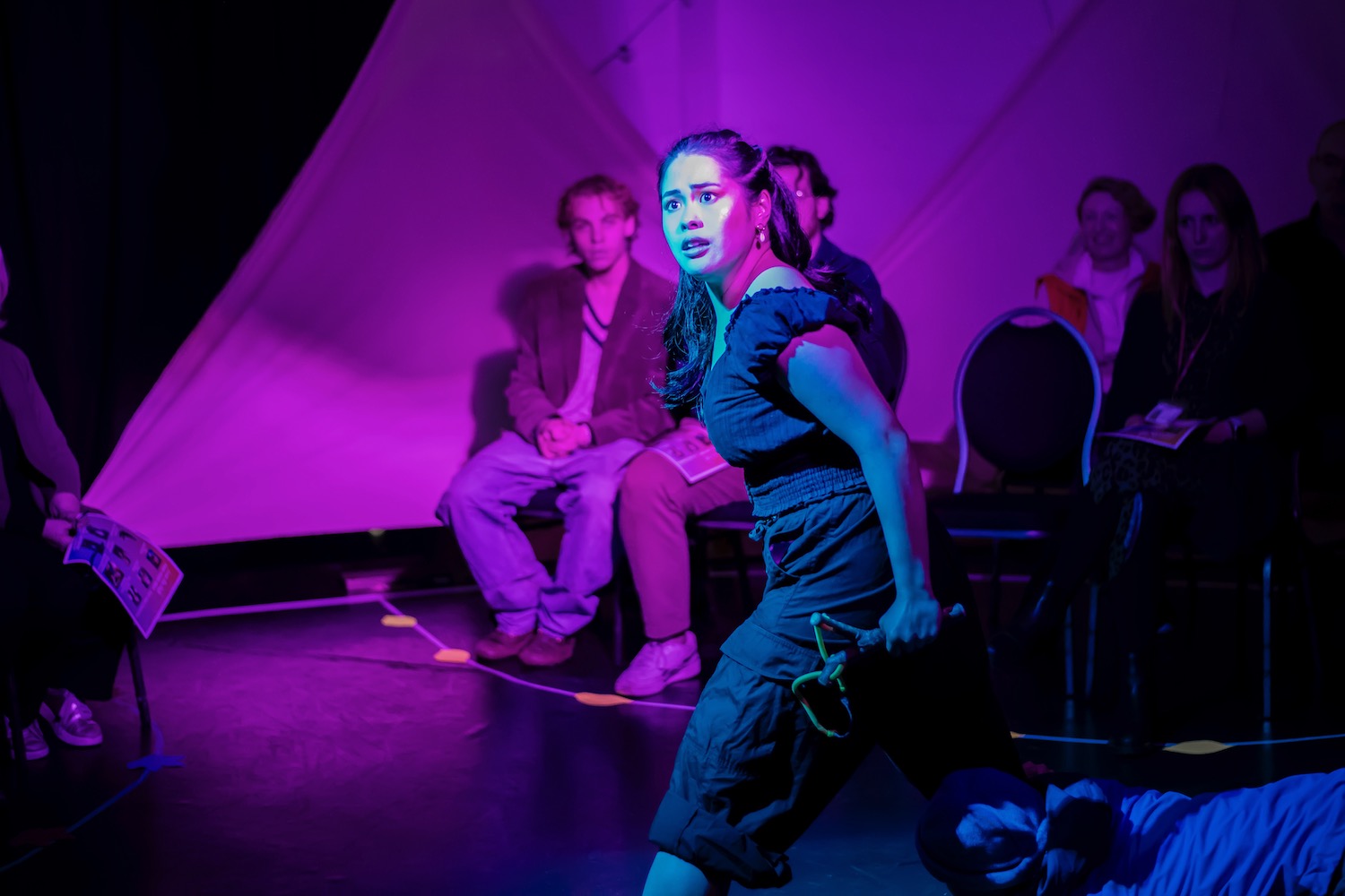 a woman performing in Lose the Path, Find Your Way. She is holding a slingshot and looking at something in the distance with a concerned expression