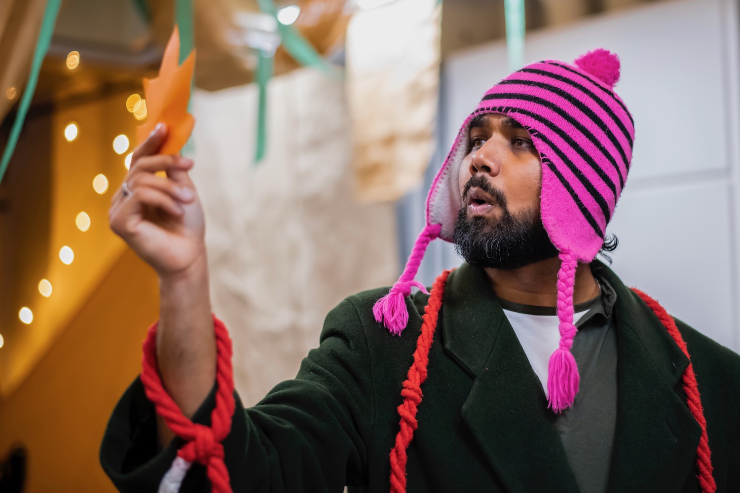 A man wearing a bright pink bobble hat is looking at an orange paper leaf reading something that is written on it