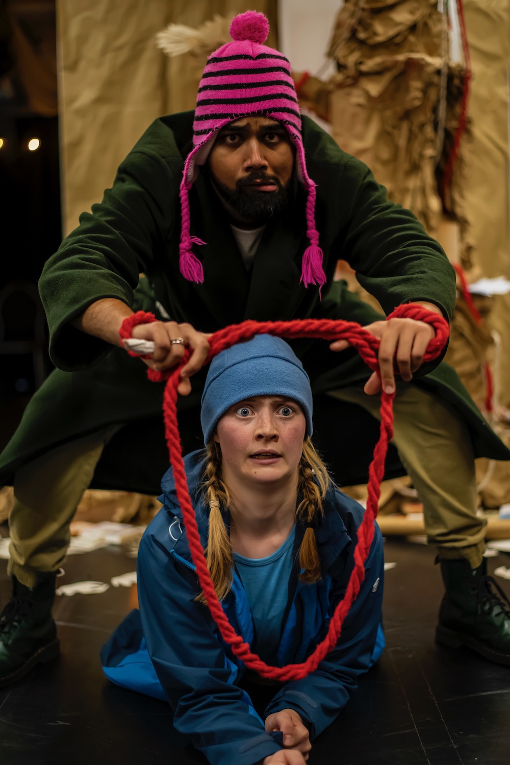 a male and female actor are peering through a piece of red rope as if it is a portal to another world and are recoiling from whatever they can see on the other side