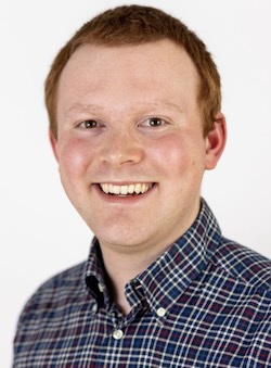 A photo of Josh, a white man, with short ginger hair, wearing a chequed shirt, smiling at the camera