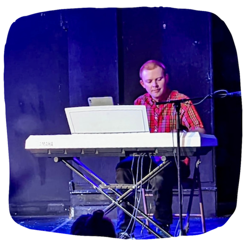 An image of Josh sitting behind a white keyboard, performing on stage. He is wearing a red shirt and there is blue lighting
